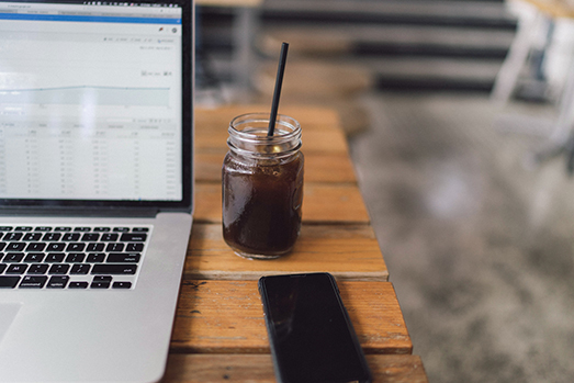 mac laptop on a table next to an iced coffee and iphone